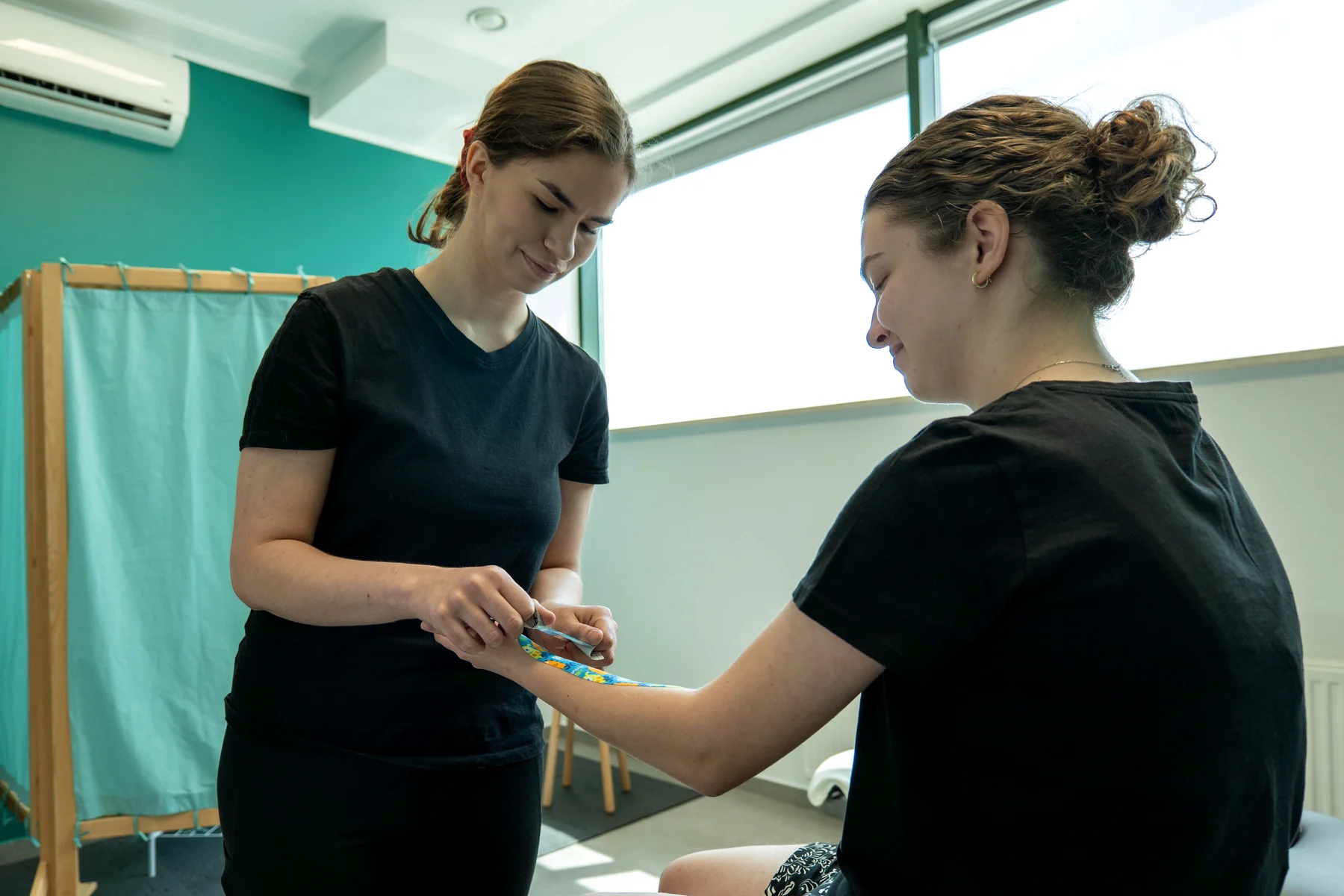 Kinesiotaping w M&M Zdrowy Masaż — fizjoterapeutka nakleja kolorową taśmę kinezjologiczną na przedramię pacjentki